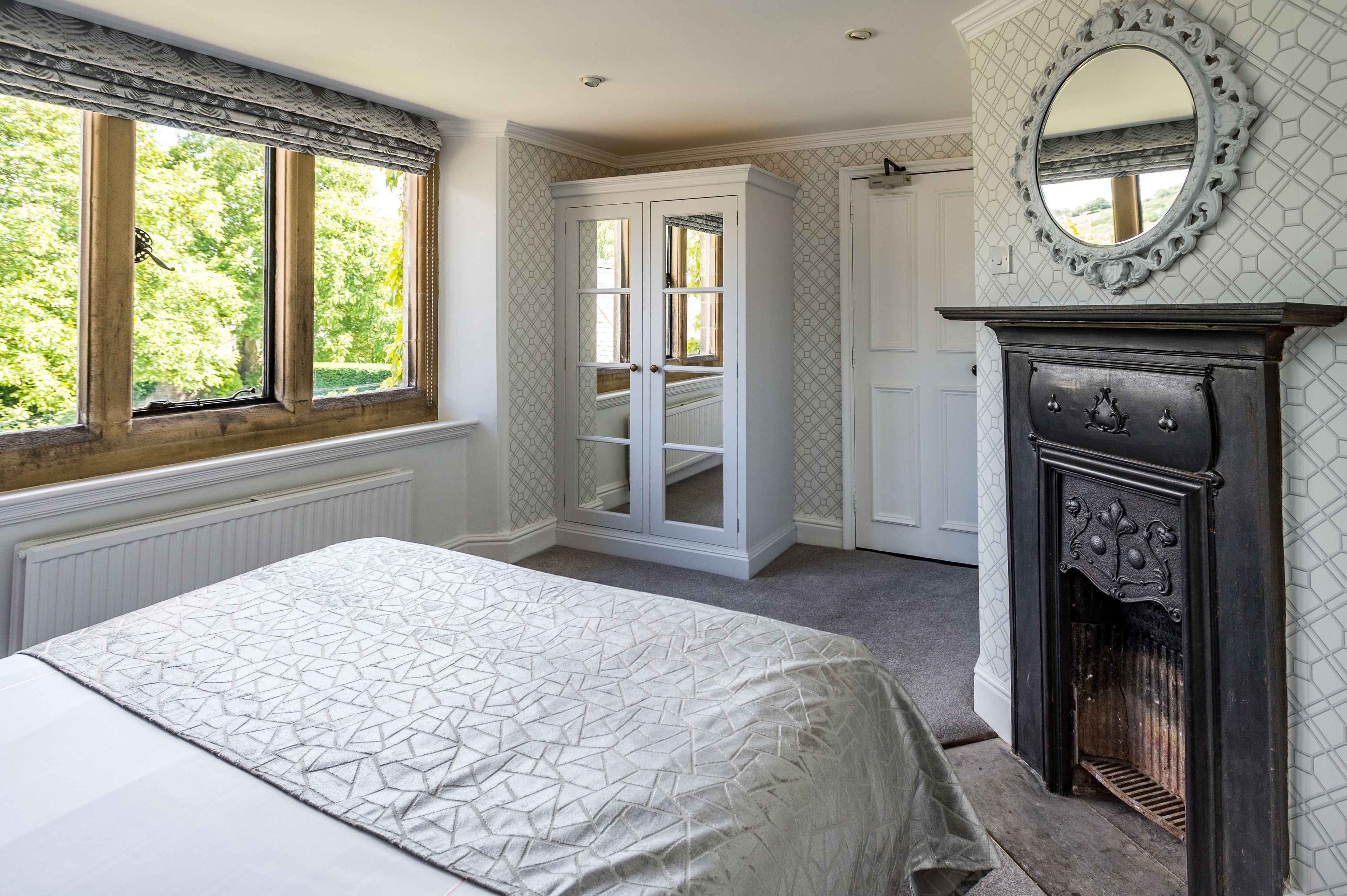A bedroom with a bed, a wardrobe, a fireplace, and a window with a view of greenery.