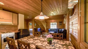 A wooden cabin interior with a dining table, chairs, a kitchen area, and a staircase leading to a living area.