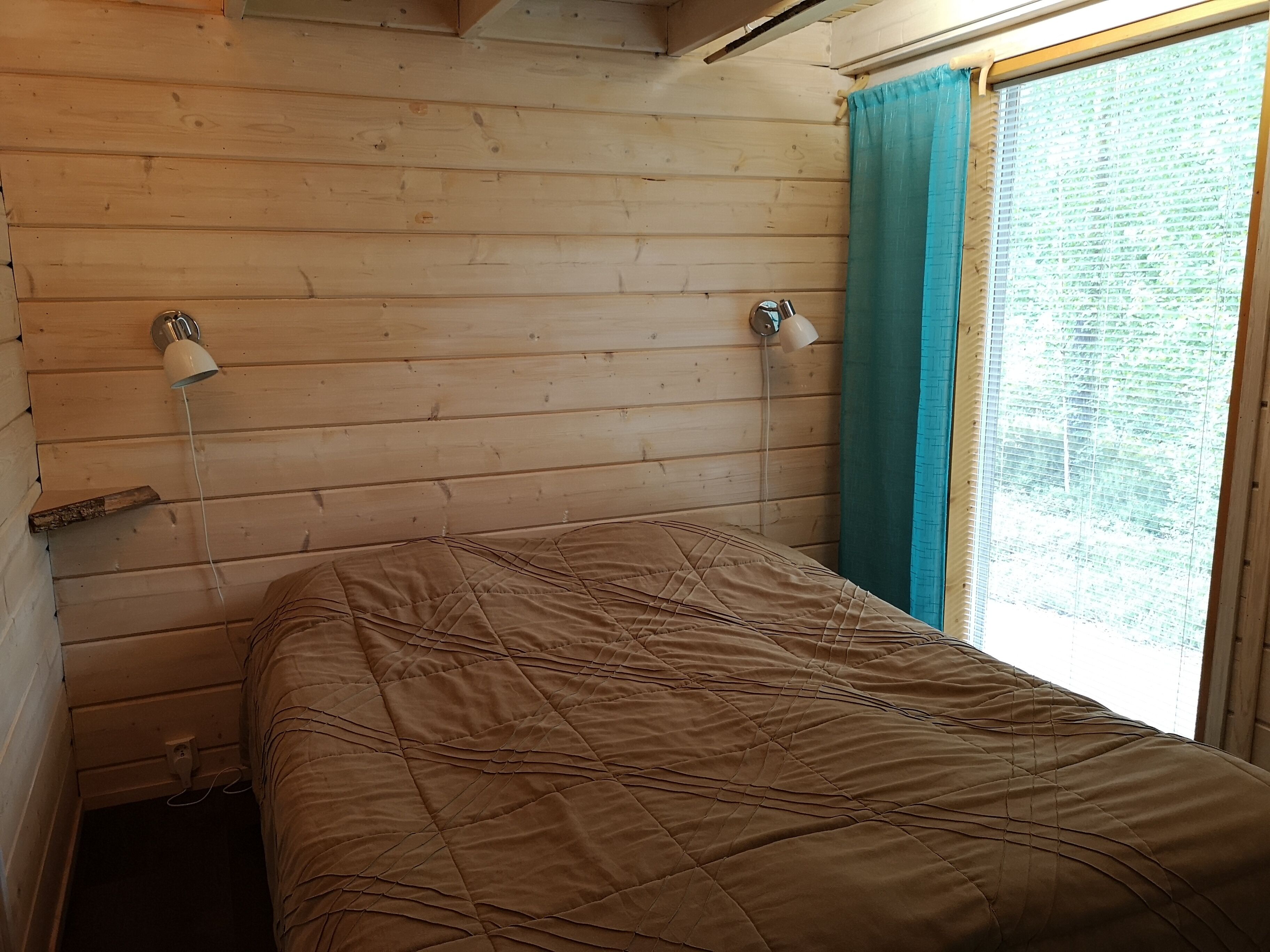 A wooden cabin room with a bed, two wall-mounted lamps, and a window with curtains.