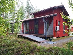Punainen mökki, jossa on katettu kuisti, ramppi ja pieni ulkoalue, jossa on matto ja harja.