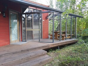 Puinen terassi, jossa on lasiseinät, punainen rakennus, jonka päällä on numero «2», ja piknikpöytä.