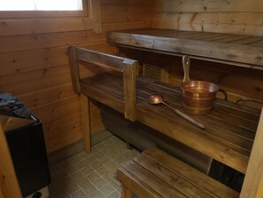 Puinen mökki, jonka sisätiloissa on penkki, pöytä ja kuparinen kattila.