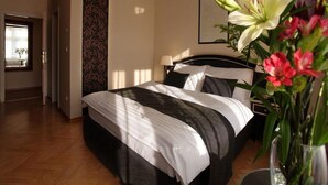 A hotel room with a large bed, a nightstand, a lamp, a window with blinds, and a wall with a patterned wallpaper. - Vesta Domus (Rome)