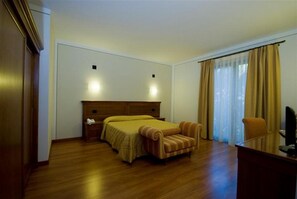A hotel room with a wooden bed, a small armchair, a desk with a chair, and a window with curtains. - Vesta Domus (Rome)