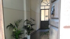 An indoor corridor with a tiled floor, white walls, and a wooden door with a brass handle. There are potted plants along the walls. - Vesta Domus (Rome)