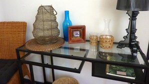 Una mesa con cubierta de vidrio, muebles de mimbre, un florero azul, un cuadro enmarcado y una lámpara negra.