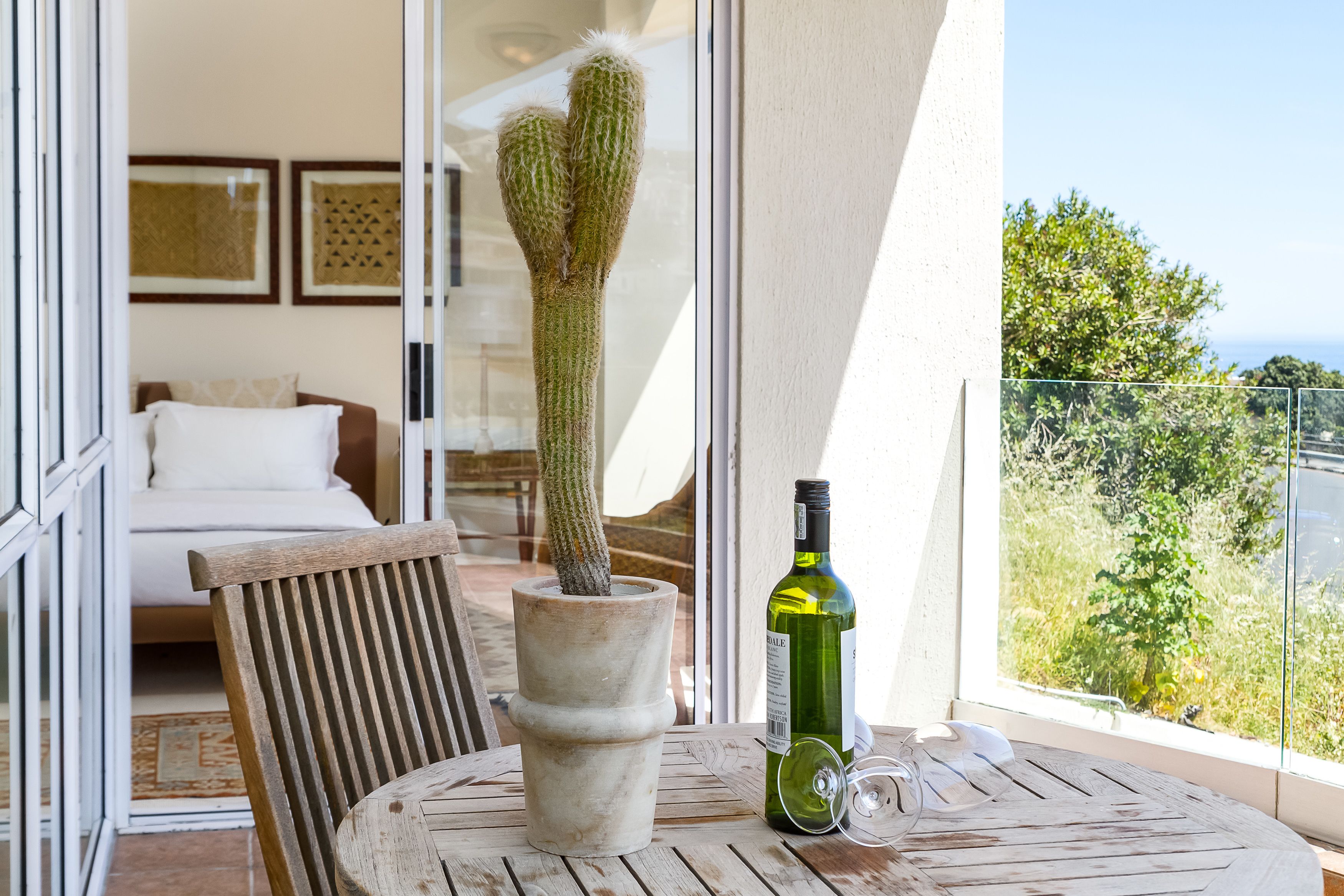 A cactus in a pot on a balcony table, a bottle of wine, and a view of greenery and a building.