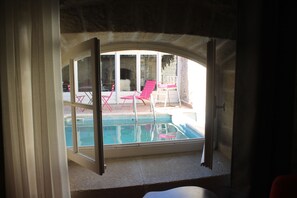 Vue depuis une chambre sur un espace extérieur avec piscine, agrémenté d’un mur en pierre, d’une chaise rouge et d’une chaise blanche.