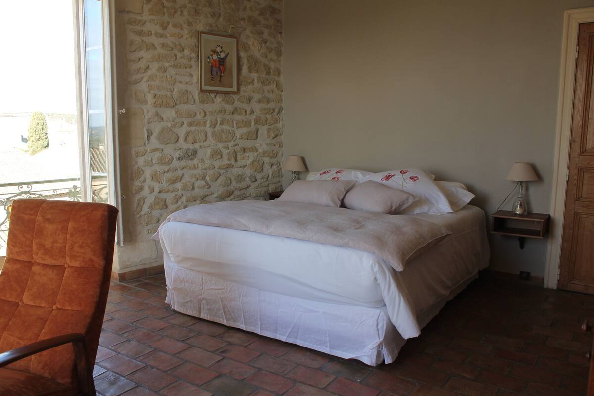 A bedroom with a stone wall, a bed with white bedding, a chair, and a lamp on a nightstand.