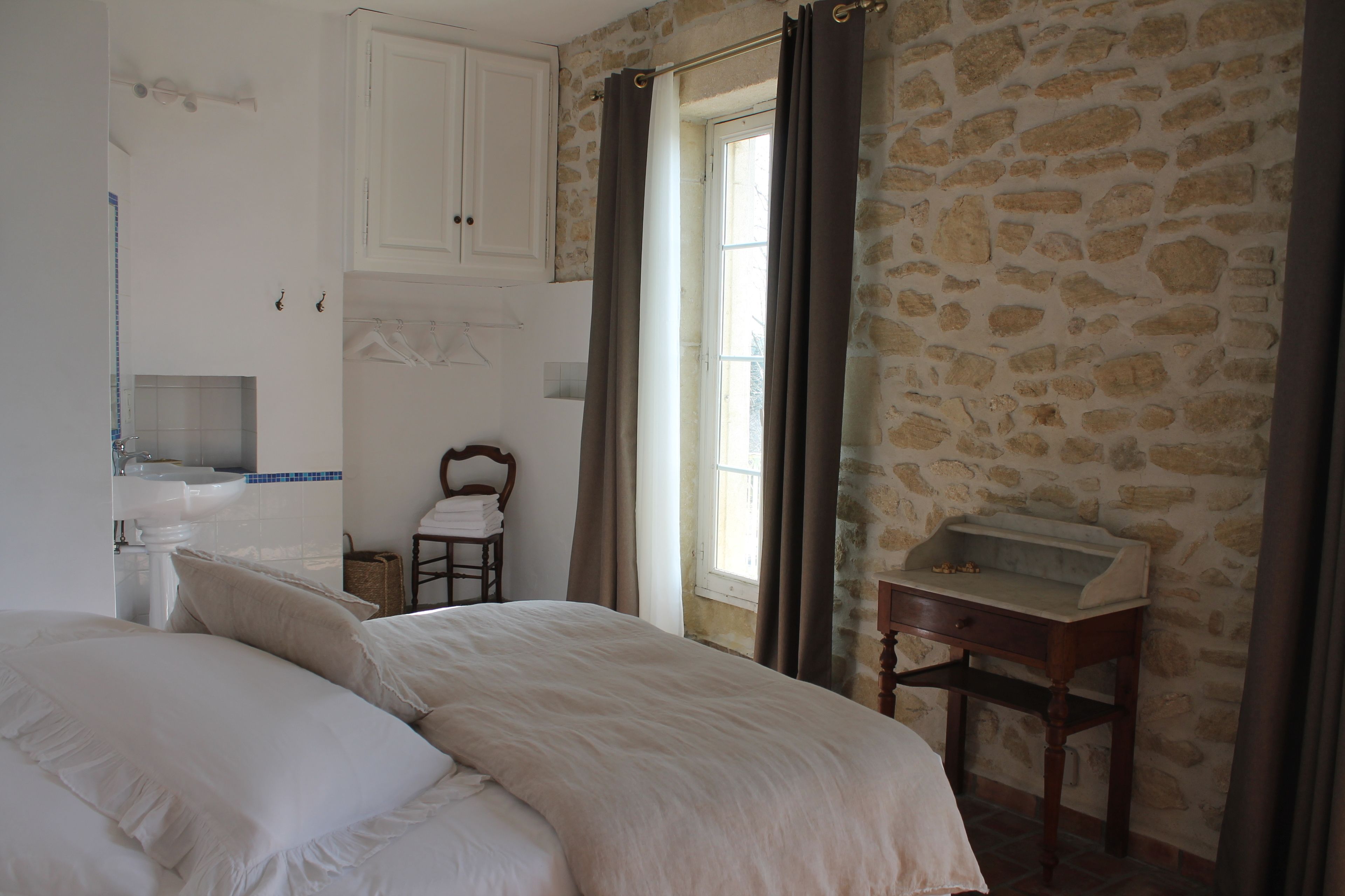 Une chambre avec un grand lit, une table de chevet en bois, un mur en pierre, une fenêtre avec des rideaux et une salle de bain équipée d’un lavabo et d’un porte-serviettes.