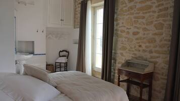 Une chambre avec un grand lit, une table de chevet en bois, un mur en pierre, une fenêtre avec des rideaux et une salle de bain équipée d’un lavabo et d’un porte-serviettes.