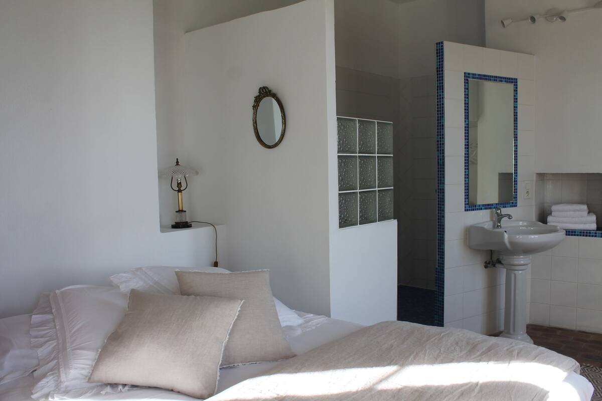 A bedroom with a bed, pillows, a bedside lamp, a mirror, and a bathroom with a sink and tiled walls.