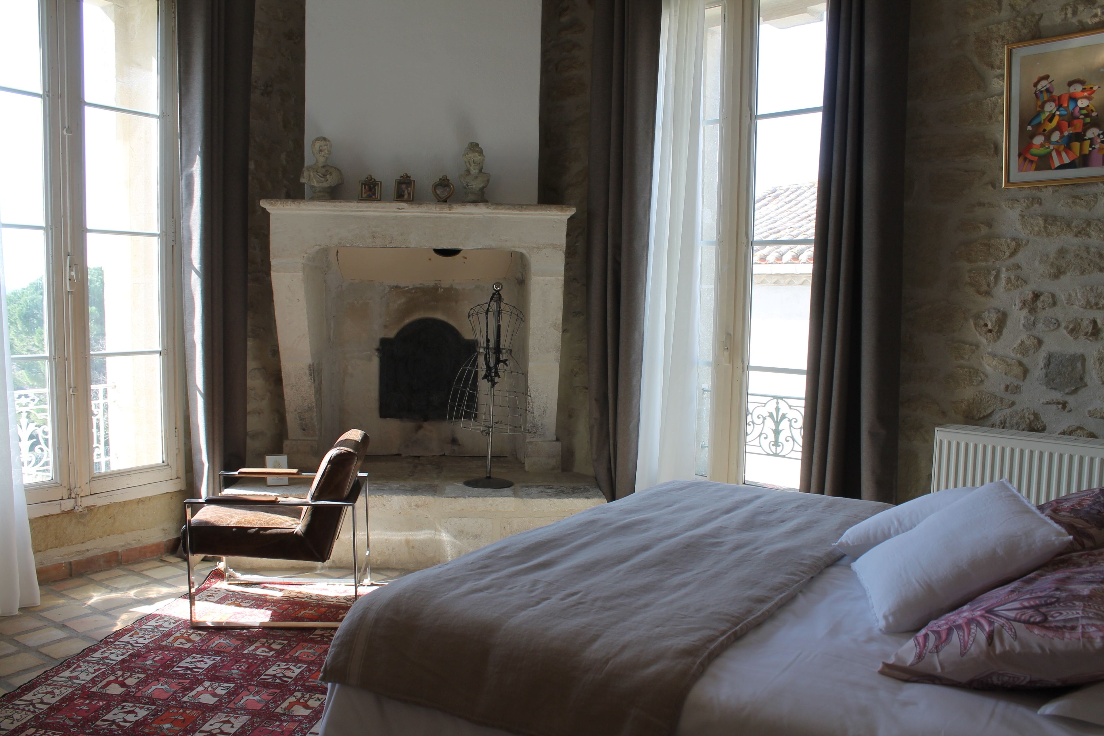 Une chambre avec un grand lit, une cheminée, un fauteuil et une vue sur l’extérieur.