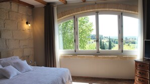 Un dormitorio con pared de piedra, un gran ventanal de arco, cama con ropa blanca y cómoda de madera.