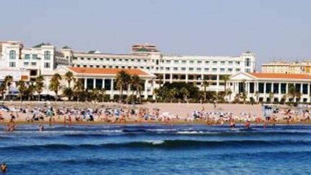Un hotel frente a la playa con varios pisos, una playa en primer plano y cielo despejado.