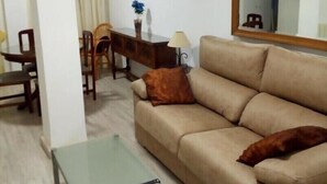 Ein Wohnzimmer mit beiger Couch, einem Glastisch, einem Holznachttisch mit Lampe, einem Sessel und einem Esstisch mit Stühlen.
