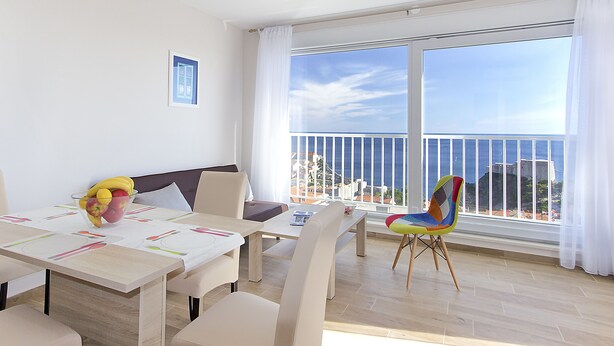 A dining area with a table set for four, a colorful chair, and a view of the sea through large windows. - Villa Ragusa Palace-Funjestra (Dubrovnik)