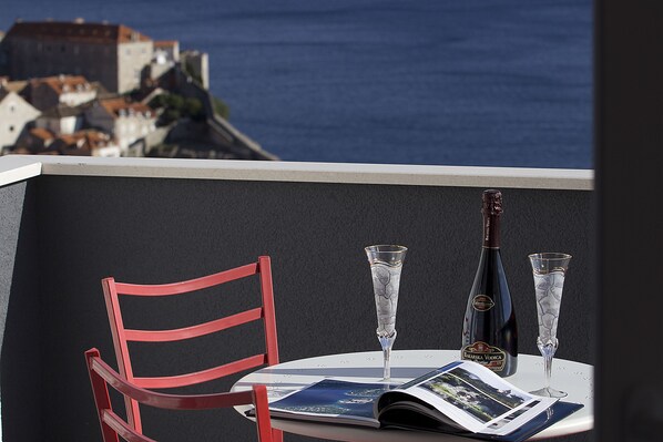 A table with a magazine, a bottle, and two glasses on a balcony overlooking a coastal town.