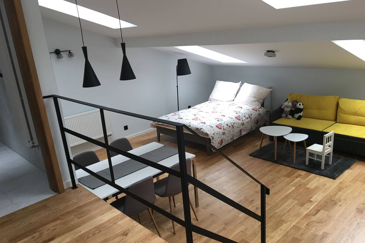 Ein moderner, gut beleuchteter Raum mit einem Bett, einem gelben Sofa, einem Esstisch mit Stühlen und einem kleinen Tisch für zwei Personen.