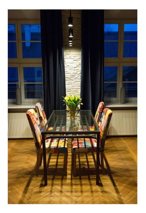 A dining area with a glass table surrounded by colorful chairs, a vase with flowers, and a stone wall behind. - Oldsmobil-Aparts (Kraków)