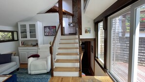 A spacious living area with a wooden staircase, a white sofa, a blue rug, and a large window offering a view of the outdoors.