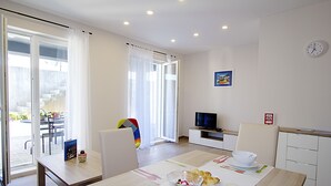 A modern dining area with a table set for two, a television in the background, and a view of the outdoors through a sliding glass door. - Apartments Ragusa Palace- Dardin (Dubrovnik)