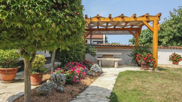 Ein Garten mit einer Pergola, Kübelpflanzen, einem Kiesweg und einer Steinsitzbank.
