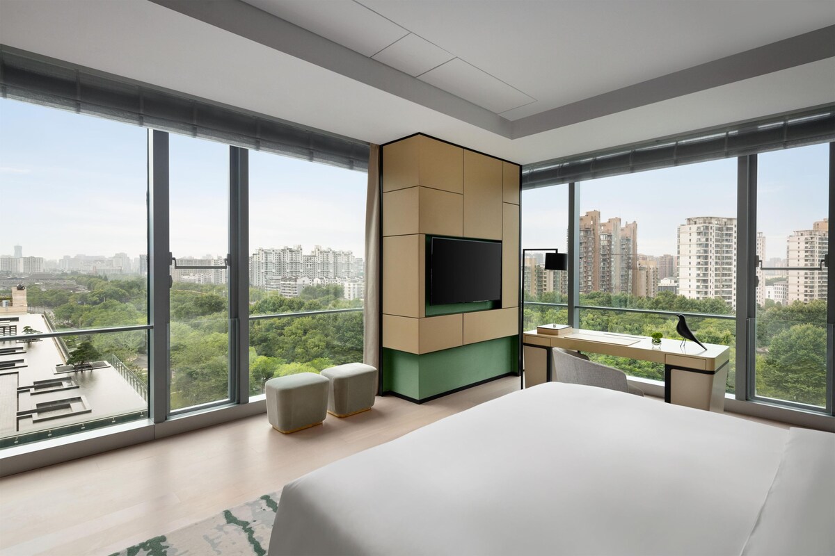 A modern hotel room with a large bed, a desk, and a TV. The room has large windows offering a city view.