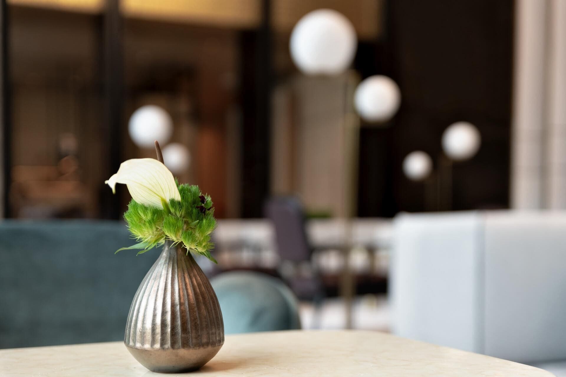 En metallvas med en vit blomma och grönt bladverk placerad på ett bord i ett modernt inrett rum.