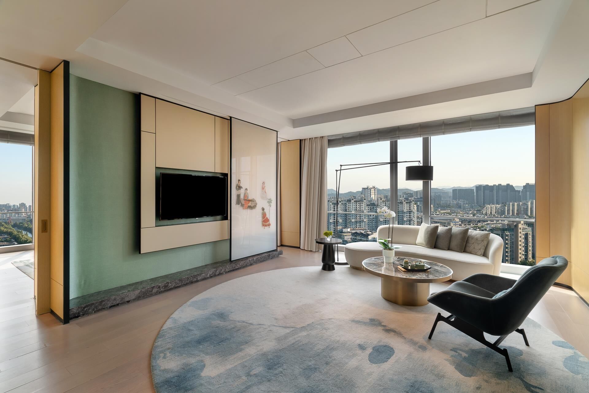 A modern living room with a flat-screen TV mounted on the wall, a round coffee table, a sofa, and a large window offering a city view.