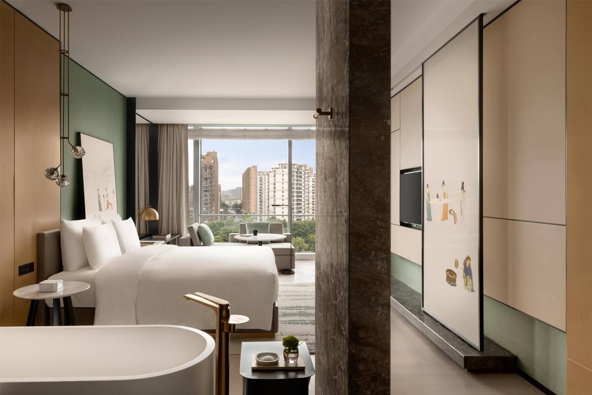 Una habitación de hotel moderna con una cama grande, una zona de estar con sofá y sillones, una bañera y vistas a la ciudad a través de amplias ventanas.