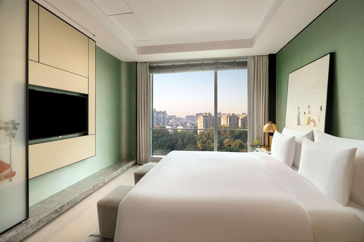 A modern hotel room with a large bed, a flat-screen TV, a city view, and a painting on the wall.