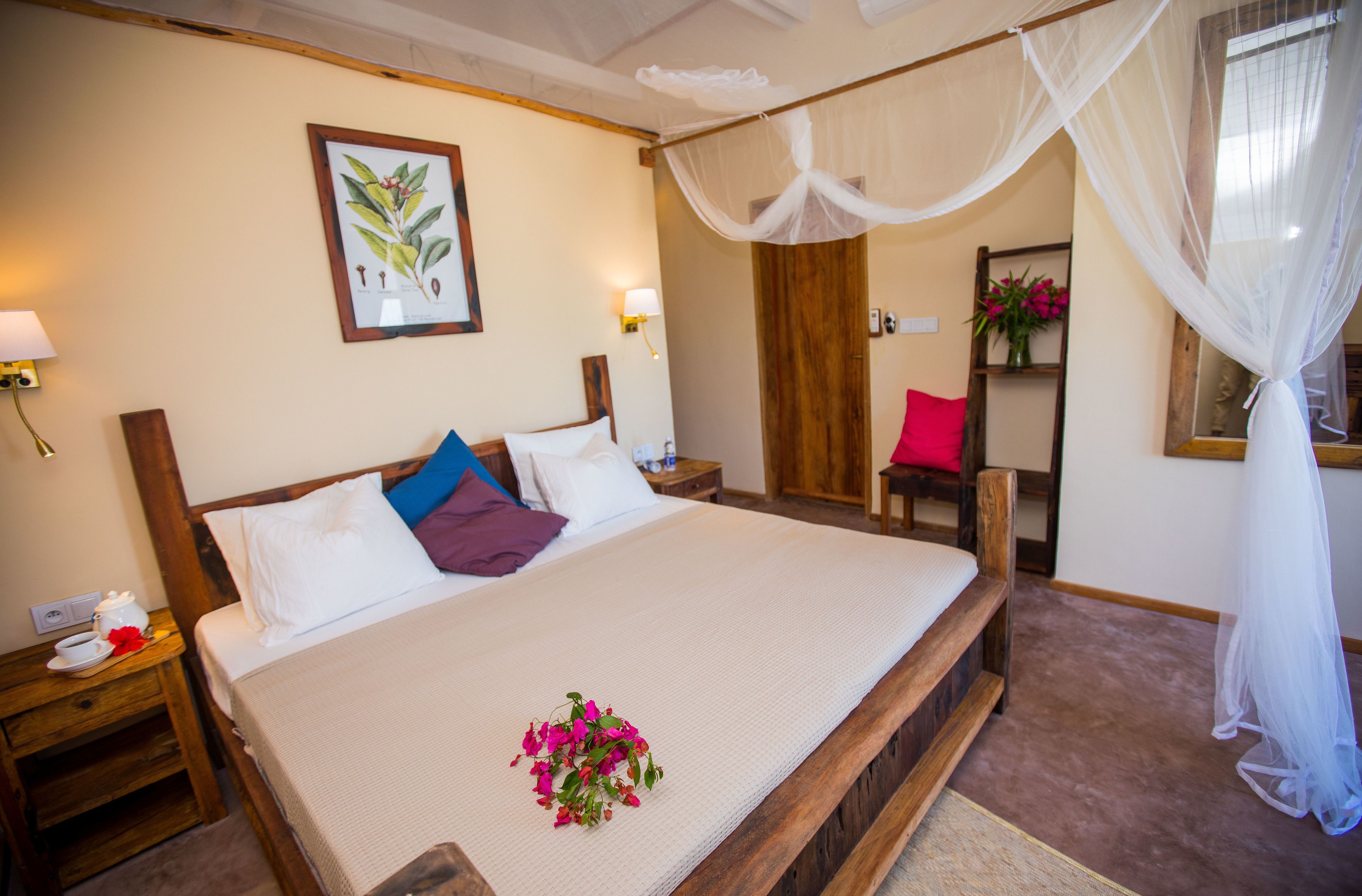 A neatly made bed with a white comforter and a bouquet of flowers. There is a wooden headboard, a nightstand with a lamp, and a wooden chair.