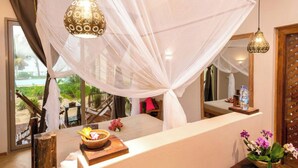 A room with a bed under a canopy, a bedside table with a bowl and flowers, and a view of the beach through large windows. - Clove Island Villas & SPA (Makunduchi)