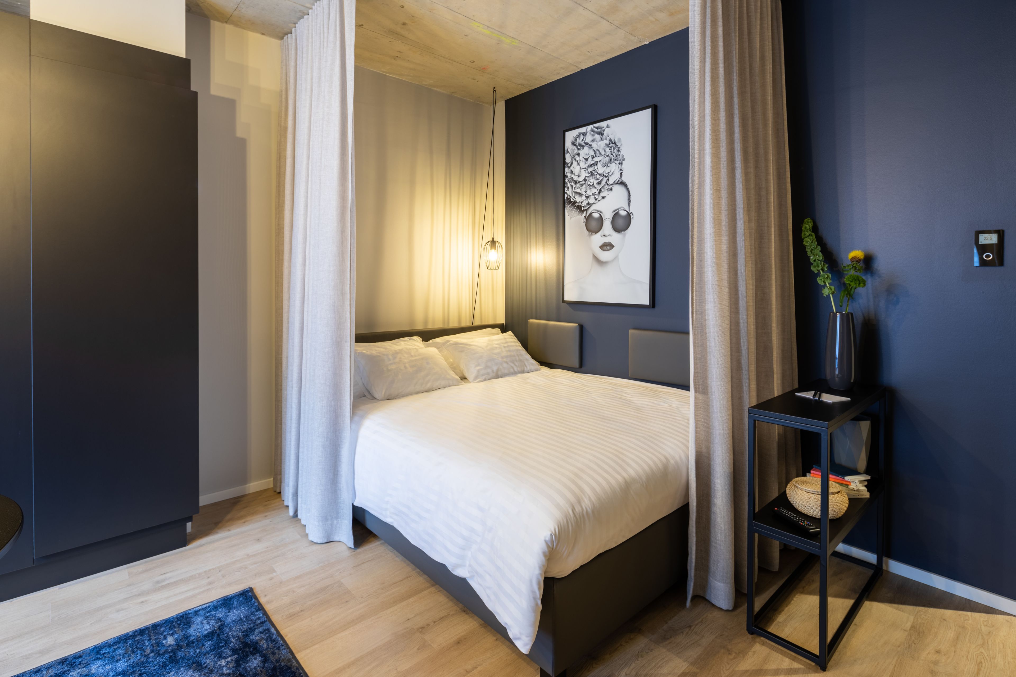 Un dormitorio moderno con cama, cortinas, una obra de arte colgada en la pared, una mesita de noche con un jarrón y suelo de madera.