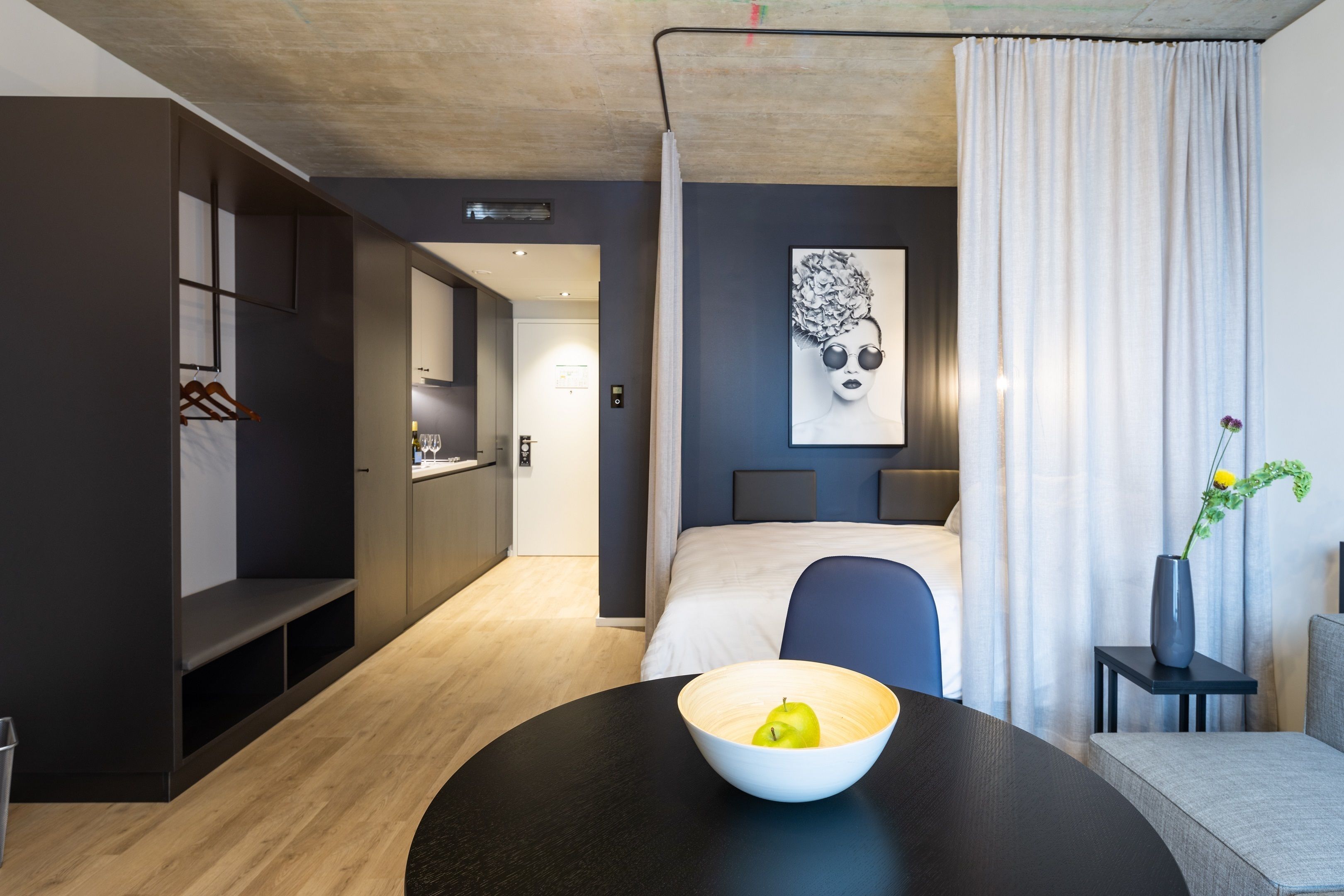 Une chambre d’hôtel moderne avec un lit, une table à manger agrémentée d’un saladier de fruits, une petite table d’appoint avec un vase, et un tableau accroché au mur.