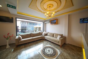 A modern hotel lobby with a beige sectional sofa, a large window, a chandelier, and a flat-screen TV mounted on the wall.