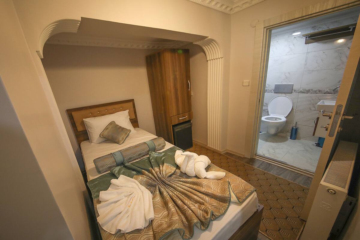 A hotel room with a bed, a wooden wardrobe, a bathroom visible through a glass door, and a patterned carpet.