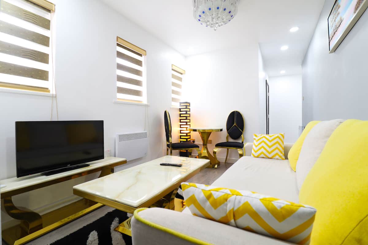 Uma sala de estar moderna com uma TV de tela plana, um sofá branco com almofadas amarelas e brancas, uma pequena mesa de jantar com cadeiras e um lustre.
