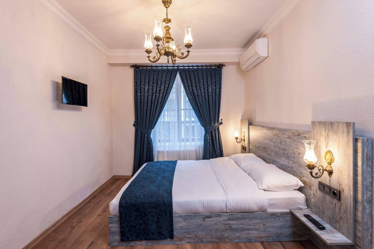 A bedroom with a bed, a chandelier, a TV, a window with curtains, and a wall-mounted light fixture.