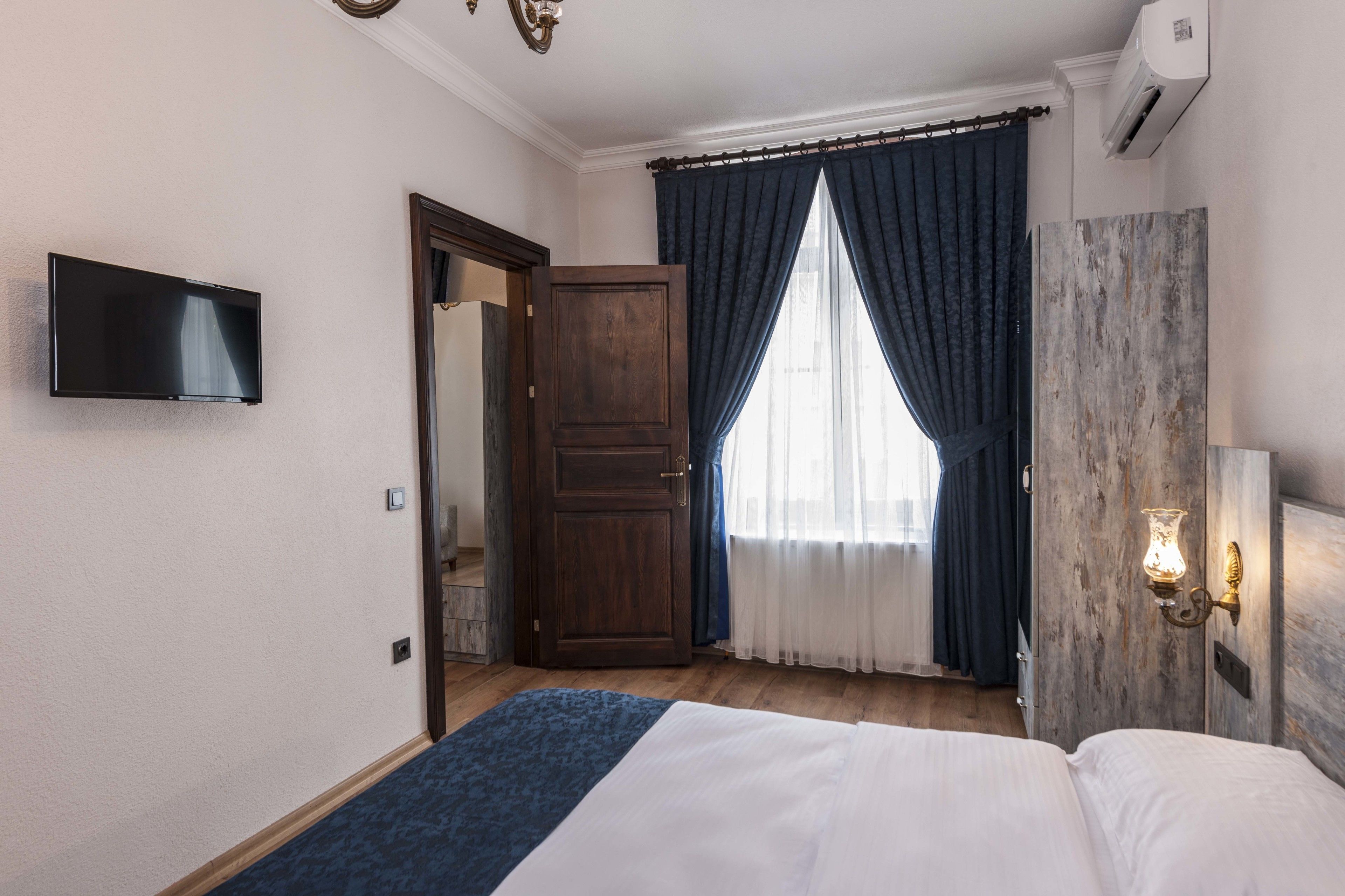 Una habitación de hotel con una cama, televisor, armario y ventana con cortinas.