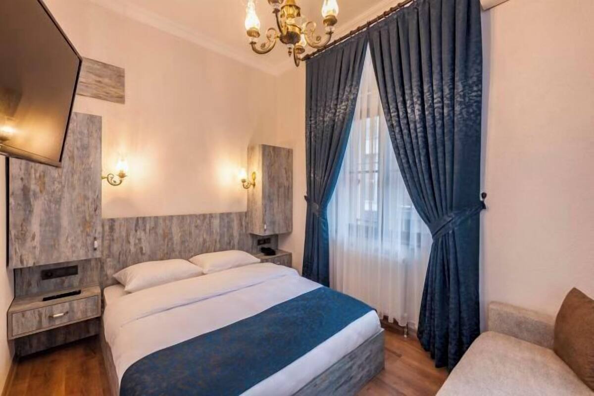 A hotel room with a bed, bedside tables, a wall-mounted TV, a chandelier, and a window with curtains.