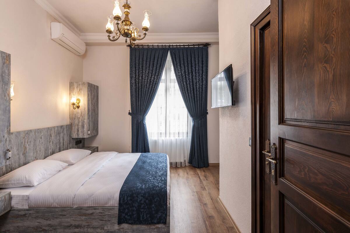 A bedroom with a bed, a wooden wardrobe, a TV mounted on the wall, and a window with curtains.