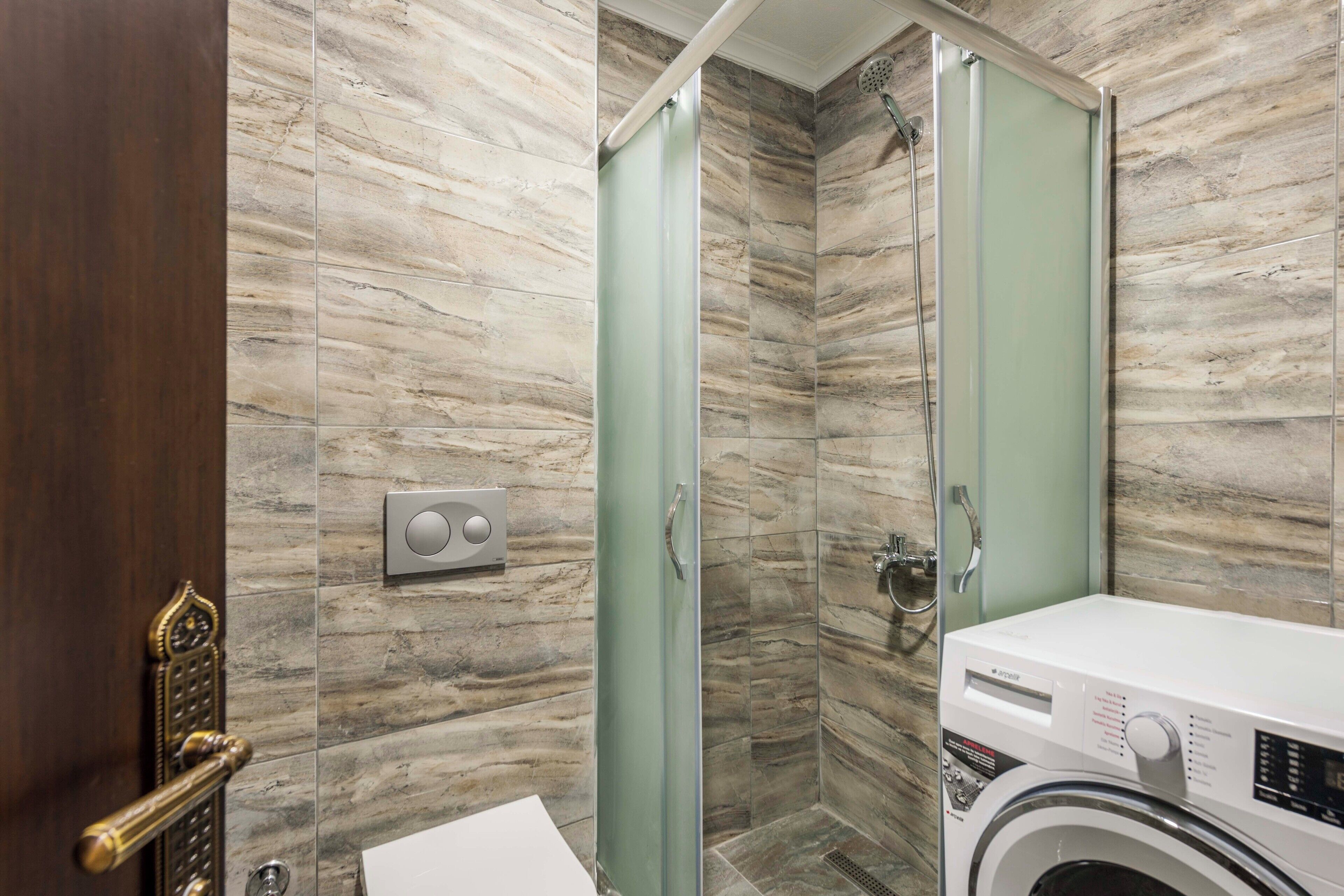 Un baño moderno con pared de mármol, cabina de ducha de vidrio, lavadora y puerta de madera con picaporte de latón.