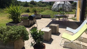 An outdoor patio with a grill, wooden planters with plants, a yellow lounge chair, a metal table with chairs, and a geodesic dome structure.