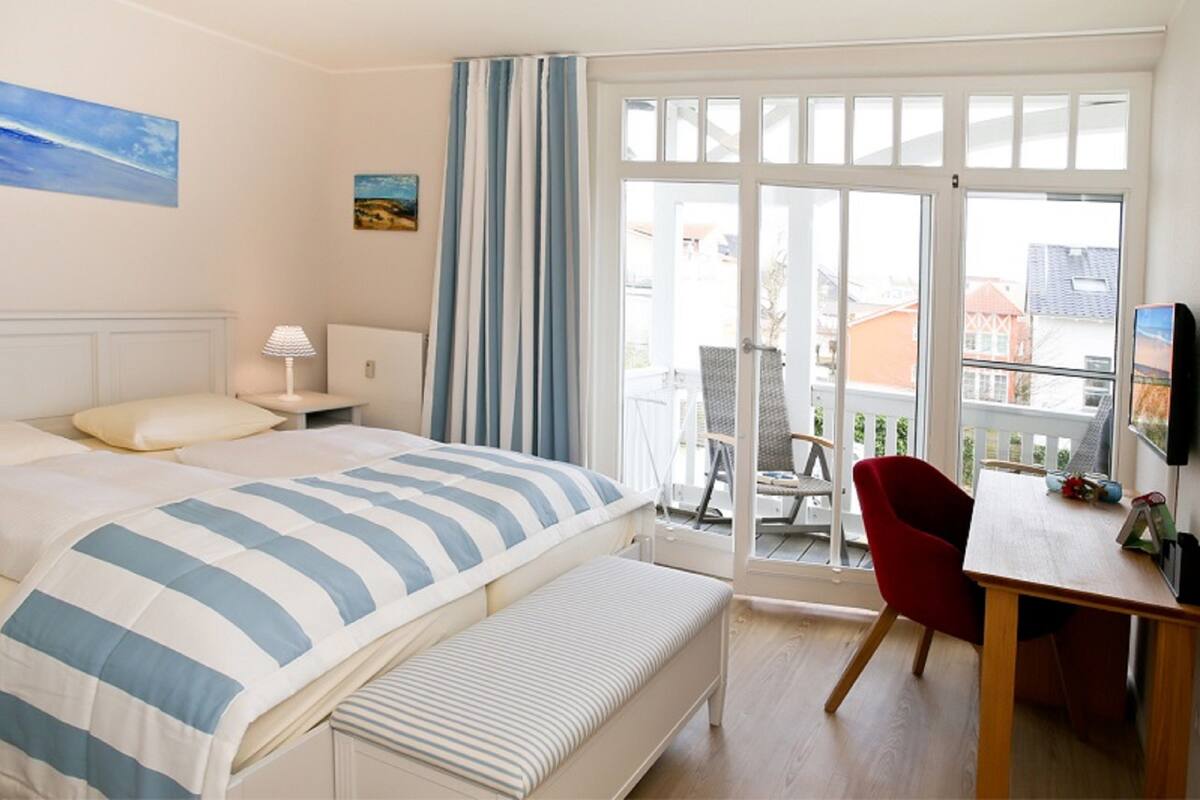 Habitación de hotel con cama, un banco, escritorio con silla, televisión y balcón con silla y mesa.