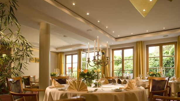 A dining area with tables set for a meal, large windows with curtains, and a chandelier. - Hotel Eisbach (Ransbach-Baumbach)