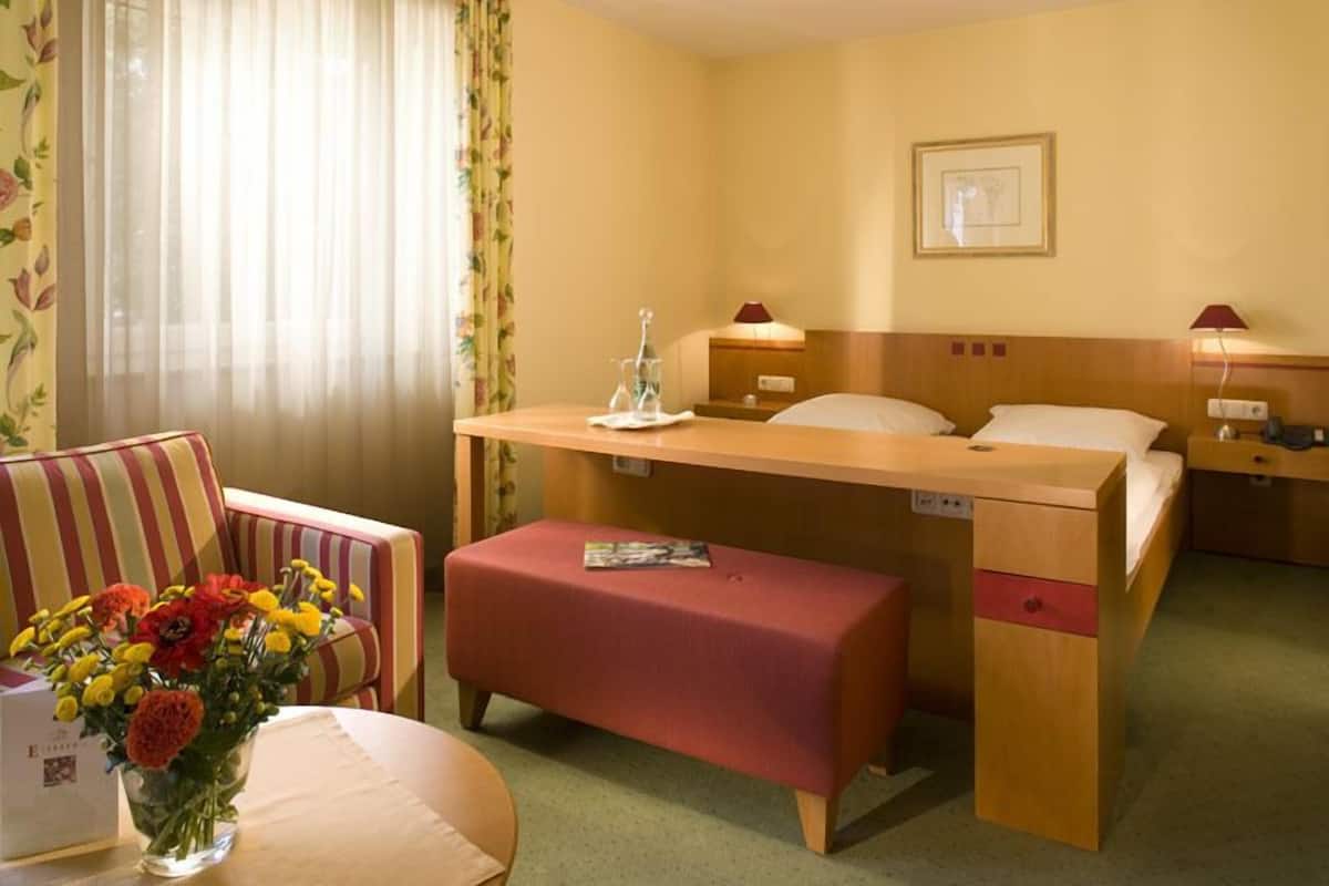 A hotel room with a bed, bedside table, lamp, and a small desk. There is a chair with a cushion and a vase of flowers on a table.