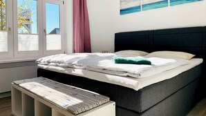 A bedroom with a bed, a bench, a window with blinds, and a view of the outside. - Apartment Hotel Rosenhof (Kuehlungsborn)
