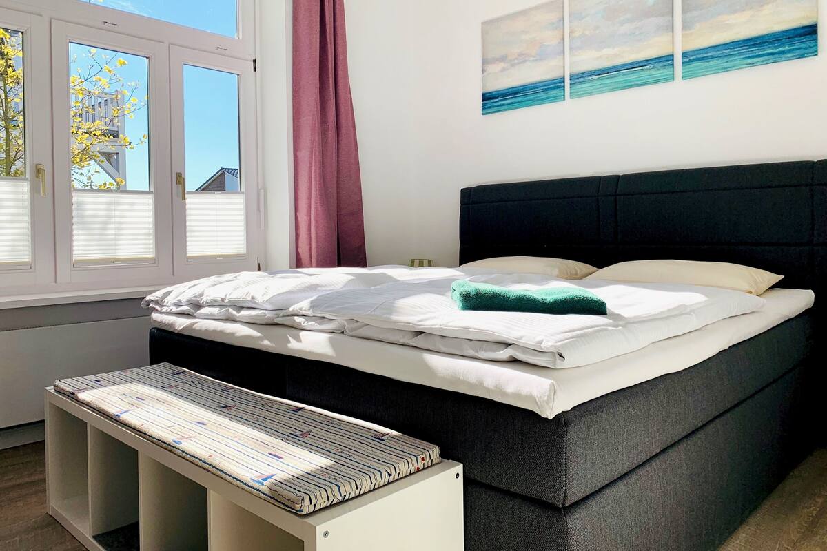 A bedroom with a bed, a bench, a window with blinds, and a view of the outside.